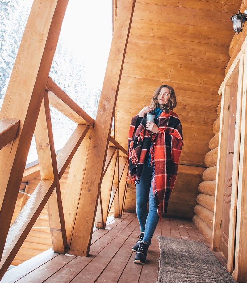 A woman in a wrap with a coffee in hand stands on the front porch of a log ski lodge in winter.