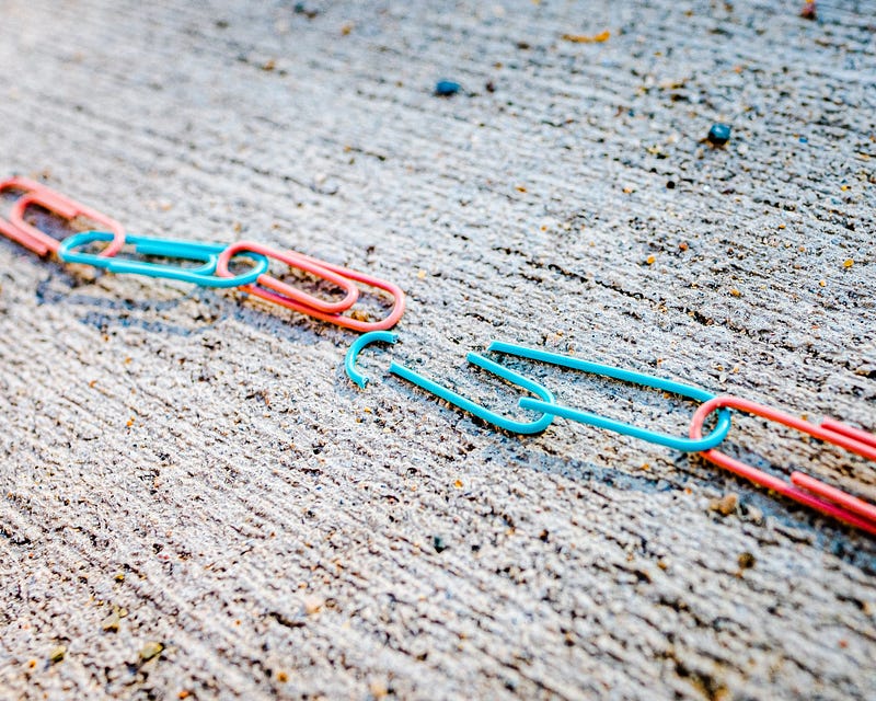A chain of pink and blue paperclips runs across a rough surface. They are broken in the middle, between a blue and pink link.