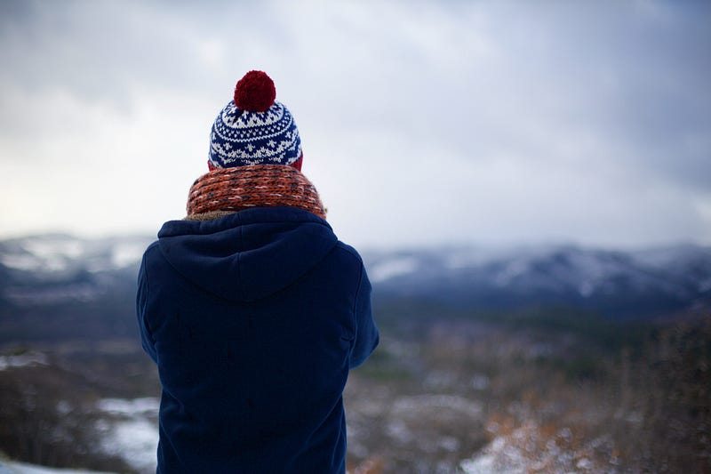 A woman in a winter coat and toque, looking down over a valley, with snowy mountains in the distance.