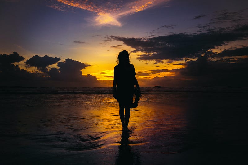 A woman walking alone on a beach at sunset.