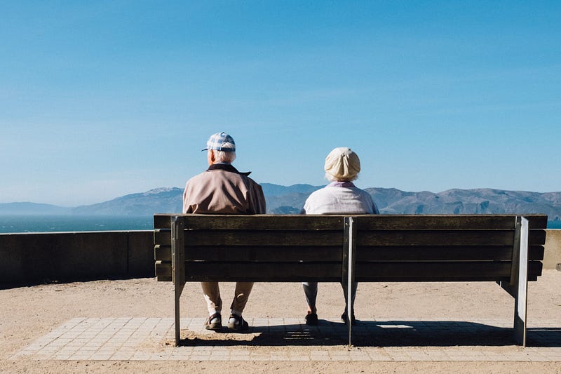 And older couple sit together on a park bench, slightly separated and looking in different directions.